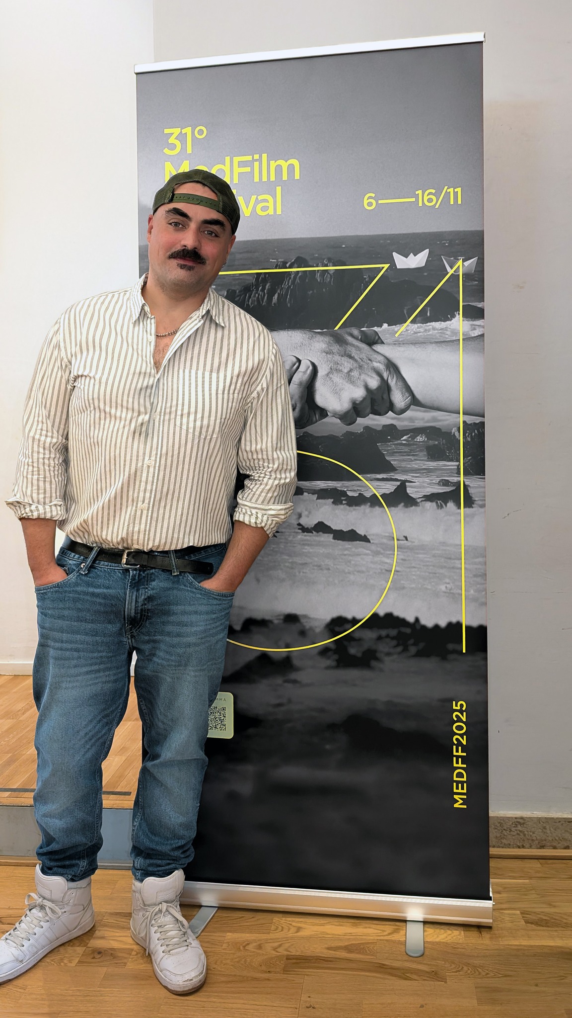 Gianluca Abbate posing in front of the poster of the 31st Med Film Festival